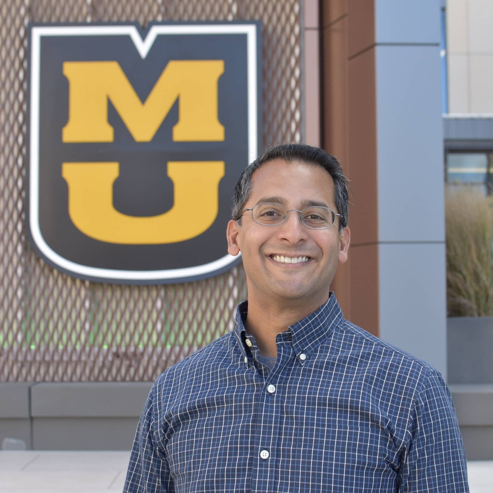 brown-skinned man with glasses, wearing a blue shirt, standing in front of a large stacked MU emblem