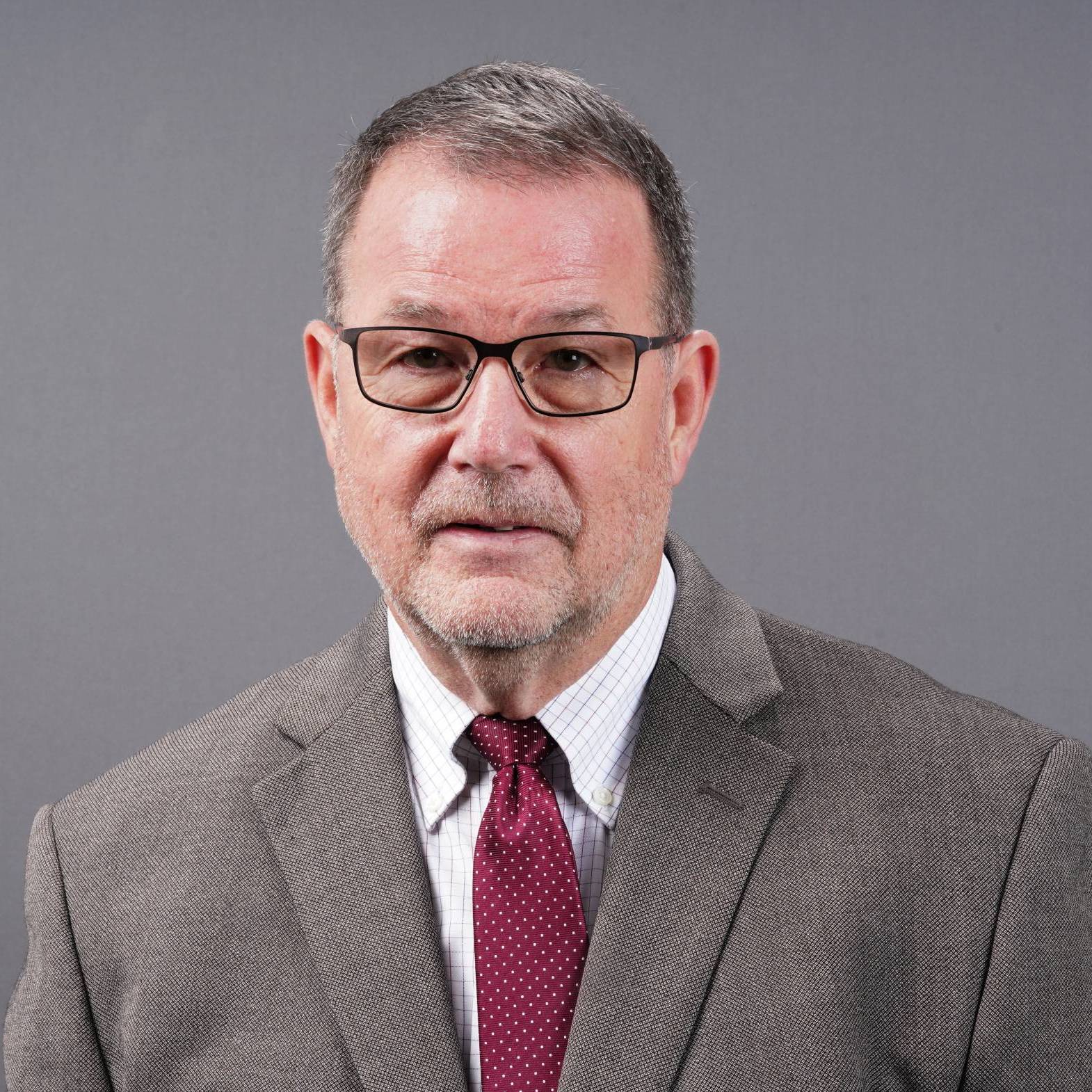 middle-age white man with brown hair and glasses wearing a brown suit jacket with a red tie
