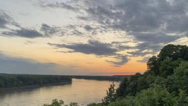 A view of the Missouri River from above. The river is bending to the left with a heavy, green treeline to the right. The sun is setting in colors of orange and yellow at the horizon.