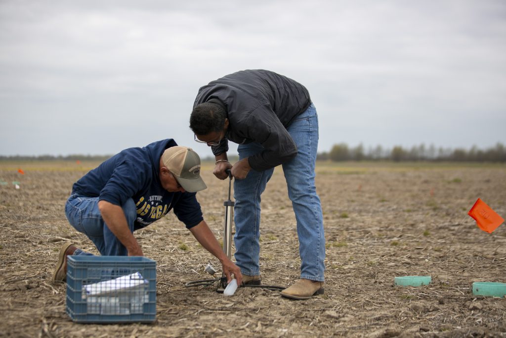 Two men in blue jeans and work boots place tubing into the ground of a harvested corn field to collect a water sample