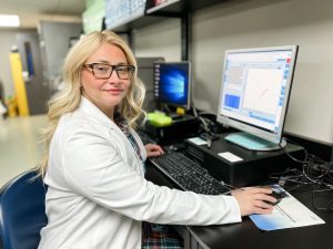 A light-skinned woman with long, blonde hair and glasses wearing a white lab coat sits at a computer.