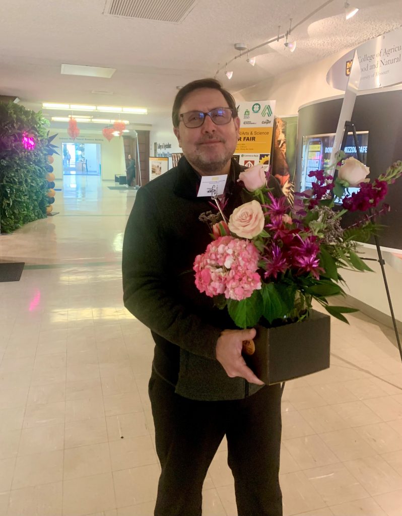 man with a dark sweater and glasses holding a flower arrangement with pink and purple flowers
