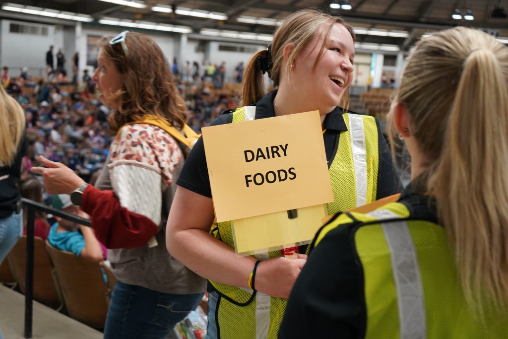 A student holds a sign reading "Dairy Foods" and leads other students through the livestock arena. 