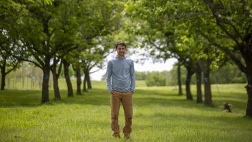 A man wearing a long-sleeve blue shirt and tan pants stands between two rows of mature trees