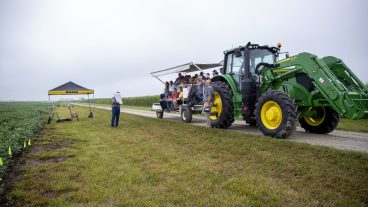 several individuals sit on risers pulled behind a tractor in a soybean field as a man explains information on a poster beneath a Mizzou branded tent