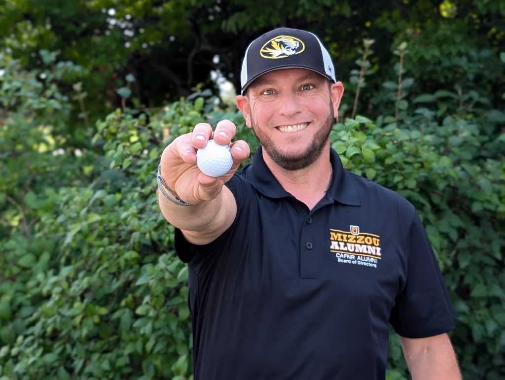 A man poses for a photo while holding a golf ball toward the camera. 