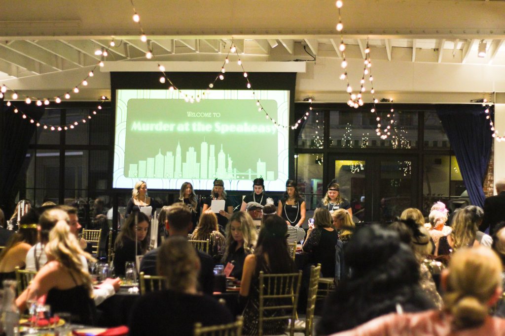 A group of students presents to a room full of people dress in 1920s attire. 