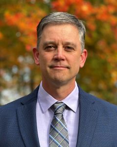 Portrait of Joel Spencer in blue suit with fall background