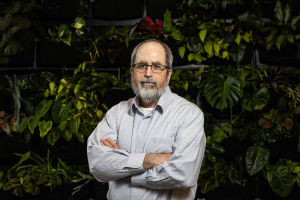 Ron Mittler stands with his arms crossed in front of a wall of green plants.