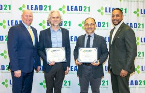 Four people stand smiling in front of white banner with the words LEAD 21 in blue and green font.