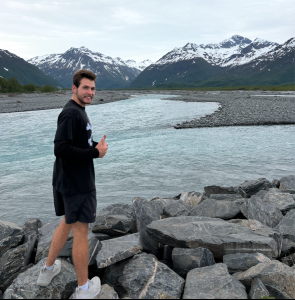 Will Lingle smiles and gives a thumbs up in front of a body of water with mountains in the background.
