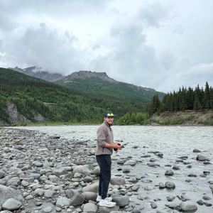 Will Lingle smiling while standing on rocks near a body of water and mountains.