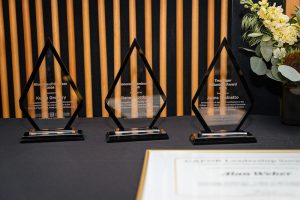 Three glass awards and an award plaque sit by flowers on a table.