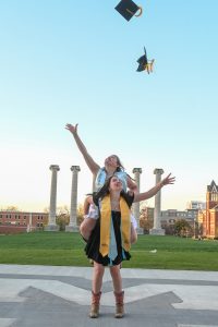 Beth Foltz carries Bell Foltz on her back while they smile and throw their graduation caps in the air in front of the Columns.