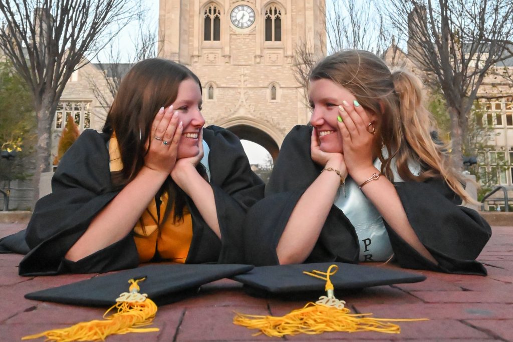 Beth and Bell Foltz smile at each other with their heads in their hands in their caps and gowns on Lowry Mall.