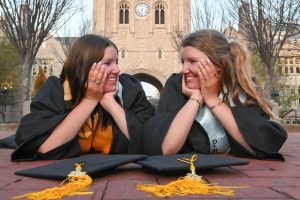 Beth and Bell Foltz smile at each other with their heads in their hands in their caps and gowns on Lowry Mall.