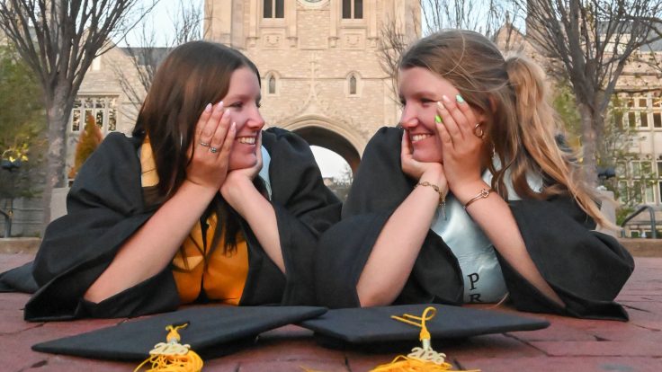 Beth and Bell Foltz smile at each other with their heads in their hands in their caps and gowns on Lowry Mall.