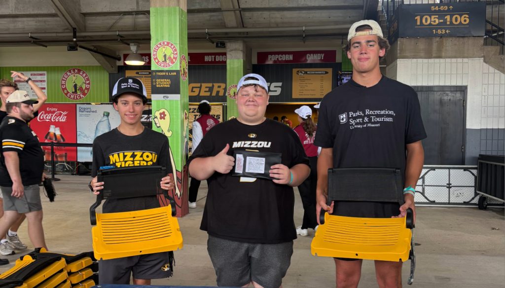Asher Skelton (middle) smiles and gives a thumbs up with two other PRST students holding bleacher chairs in Mizzou's football stadium.