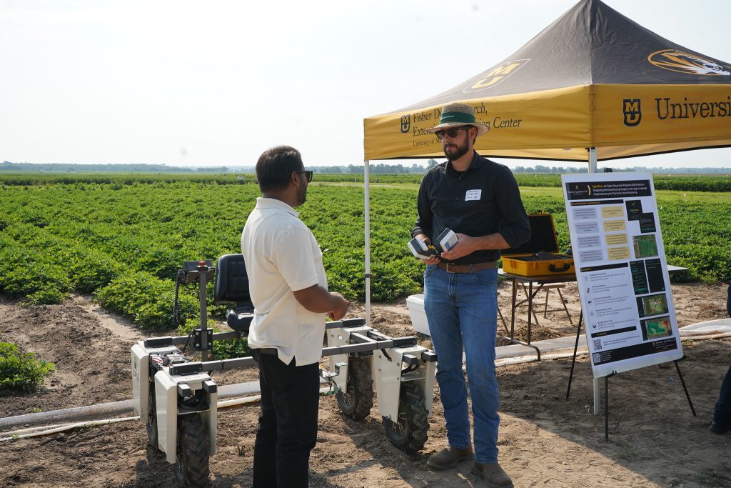 Two men chat in front of a tent in a crop field.