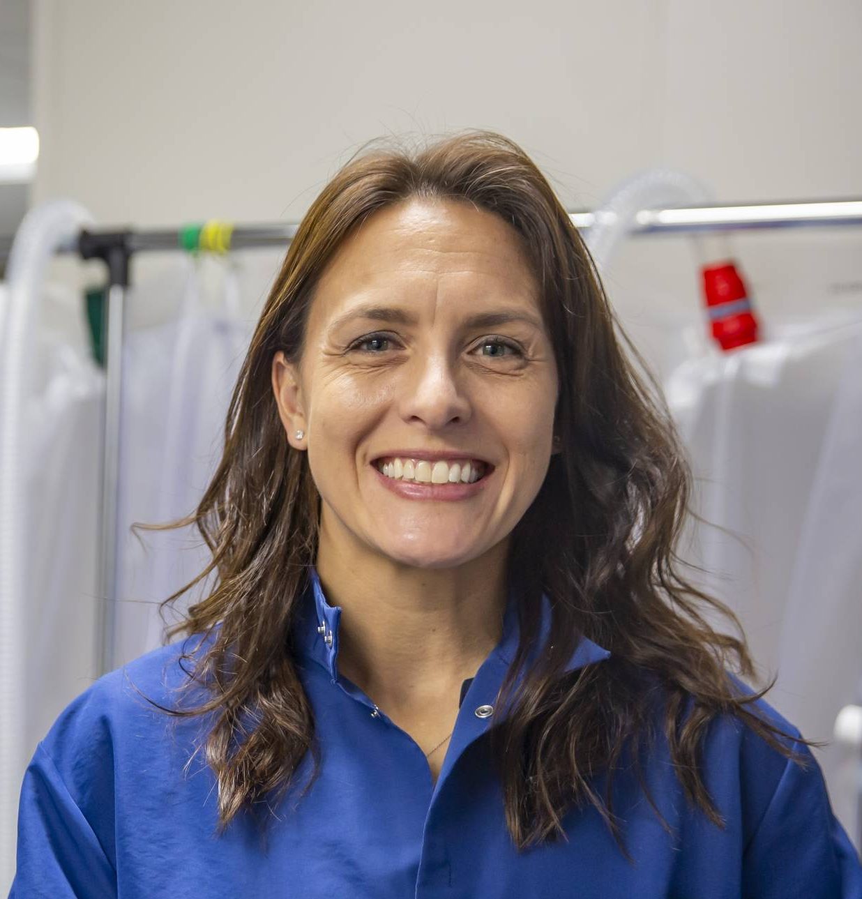 A white woman with long, brunette hair smiles while wearing a blue lab coat. Behind her are various medical equipment in a research laboratory.