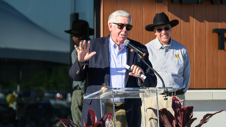 Parson talks at clear podium; Dean stands behind listening.