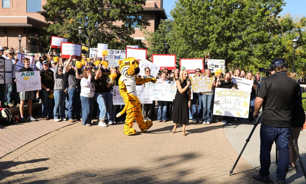 Tyne Morgan speaks into a microphone facing a camera with Truman the Tiger beside her and a crowd of students holding signs behind her.