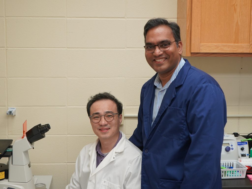 Two men pose, one standing and one sitting, in a lab.