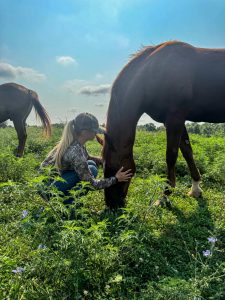 Roselynn Orr crouches down in a field while petting a grazing horse's face.