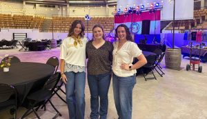 Addy Nichols, Isabella Burnine and Skylar Goodman stand together and smile in front of the Barnwarmin' setup inside Trowbridge Event Center.