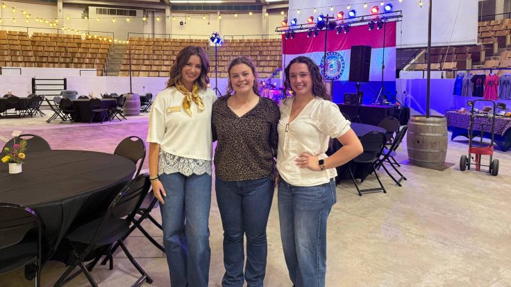 Addy Nichols, Isabella Burnine and Skylar Goodman stand together and smile in front of the Barnwarmin' setup inside Trowbridge Event Center.